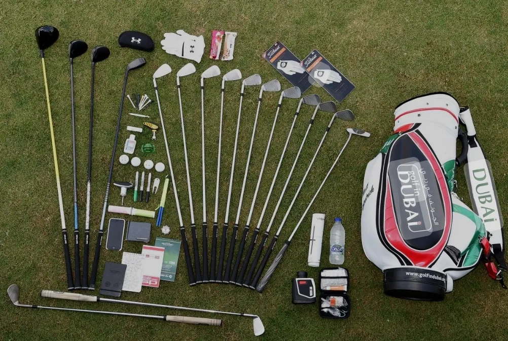 A full set of golf equipment laid out on grass: woods, irons, wedges, putter, balls, gloves, shoes, accessories and a golf bag.